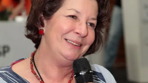 Imagem de Mariângela Furlan Haswani de cabelos curtos e castanhos, sorrindo enquanto segura um microfone. Ela usa um vestido estampado em tons de azul e branco, acessórios vermelhos, incluindo um colar e brincos. O fundo está desfocado, com pessoas e um púlpito visíveis, sugerindo um ambiente de evento ou conferência.