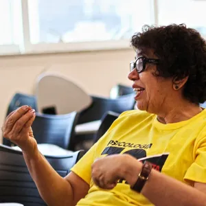 Imagem de Maria da Gória Calado de perfil, usando óculos e uma camiseta amarela com a palavra 'Psicologia' impressa. Ela está gesticulando com a mão direita à altura dos ombros, com os dedos unidos, e apresenta uma expressão facial sorridente. O fundo mostra cadeiras pretas em um ambiente interno bem iluminado.